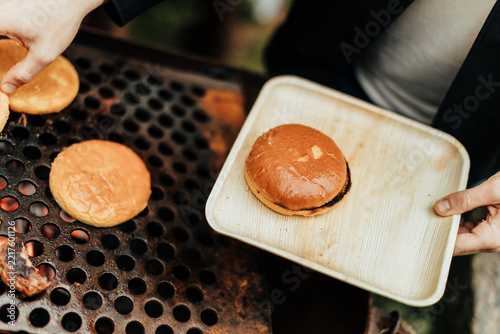 Hands hold a wooden tray with a toasted burger bun while additional buns warm on a perforated charcoal grill, creating a rustic outdoor barbecue scene