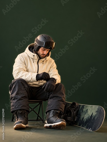 Male Snowboarder Adjusting Gloves on Folding Chair in Dark Green Studio