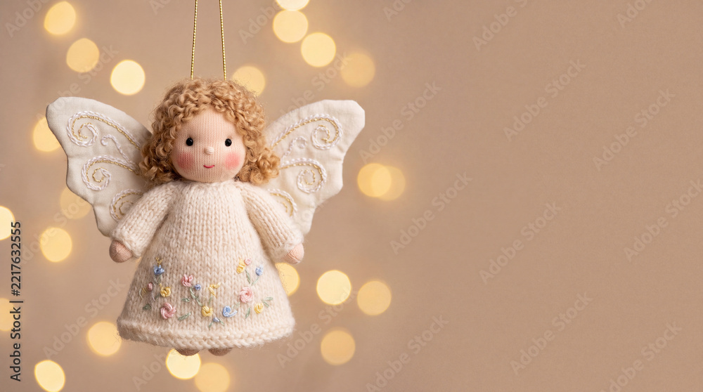 Naklejka premium Handmade angel ornament with curly hair, knitted cream dress, embroidered flowers, and white wings suspended against warm golden bokeh lights