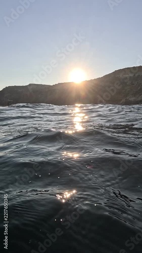 Brilliant sun setting behind rugged coastal cliffs illuminating sparkling sea waters. Low angle tracking shot.