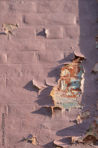 Abblättertende rosa Wandfarbe gibt freiliegendes Backsteinmauerwerk an einer verwitterten Hausfassade bei seitlichem Sonnenlicht frei; eine rustikale Detailaufnahme.