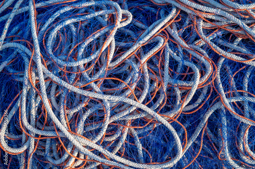 Fischernetz und Seile in Blau, Weiß und Orange liegen ungeordnet als maritime Detailaufnahme im sanften Tageslicht eines Hafens.