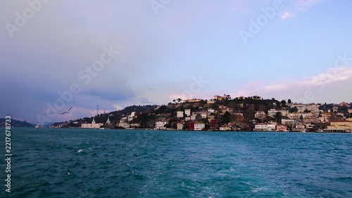 Scenic view of Istanbul and the Bosphorus Strait from a moving boat, with waterfront buildings, seagulls and a dramatic cloudy sky in the background.