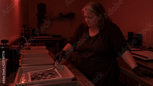 A woman wearing a dark apron leans over a table surface, carefully placing small rounded stones onto a shallow tray using tongs, the entire scene lit with deep red light