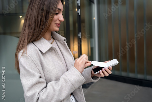 Happy young woman smiles with excitement while removing protective film from new smartphone screen outdoors, celebrating new device and modern mobile technology. Concept of joy, purchase and unboxing