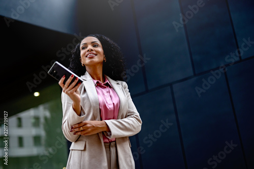 Confident professional woman holding a smartphone outside a modern office building, representing mobile technology, business communication and contemporary work