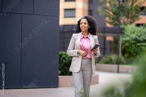 Smiling professional woman walking with a smartphone through a modern business district, representing commuting, mobile work, career confidence and urban professional life