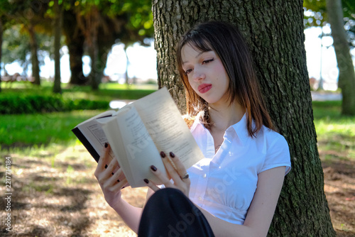 Beautiful Young Woman Reading Book While Leaning Against Tree Trunk in Park, Relaxing Girl Enjoying Literature in Green Nature Footage, Lifestyle Outdoor Leisure Concept Video, Peaceful Reading Scene