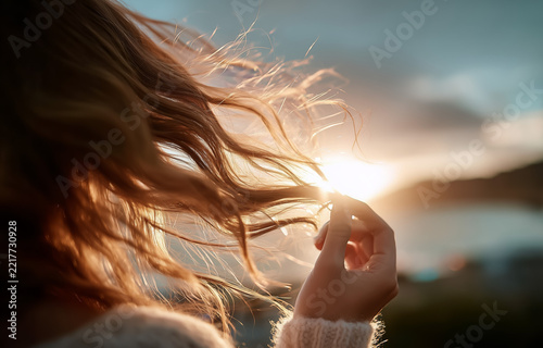 Woman touching windblown hair beside water in low sunlight