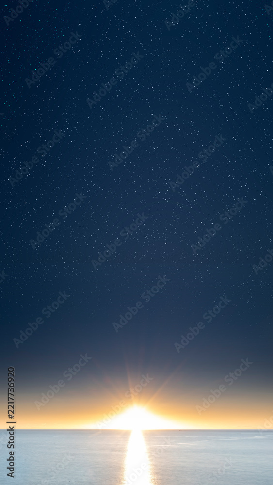 Fototapeta premium Sunrise or sunset over a calm sea under a starry night sky with bright sunburst and reflection on the water. Nature, natural or quotes background
