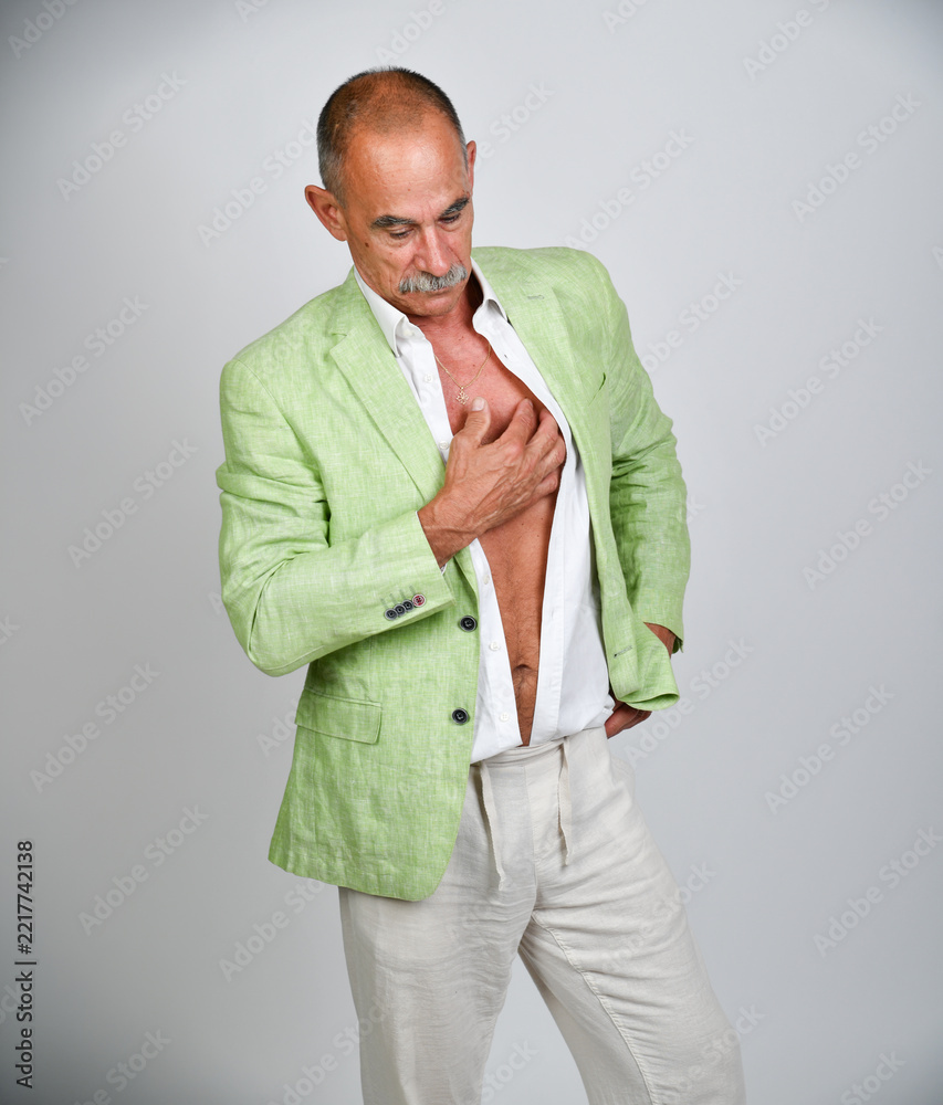 Naklejka premium Selbstbewusster älterer Mann im hellgrünen Sakko mit offenem weißen Hemd und Hand auf der Brust. Best Ager, Fashion, sommerliche Eleganz, Persönlichkeit, Lifestyle und moderne Studiofotografie.