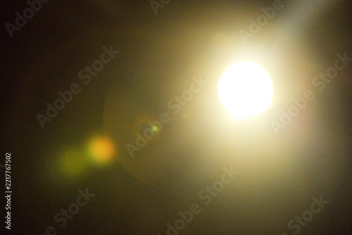 Bright circular light glowing through darkness with warm atmospheric haze and colorful lens flare, abstract illumination photograph with minimal low detail background and optical artifacts