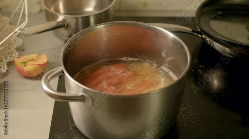 Close-up of sausages and sliced onions simmering in stainless steel pot on stovetop, with steam and sliced apple on kitchen counter.