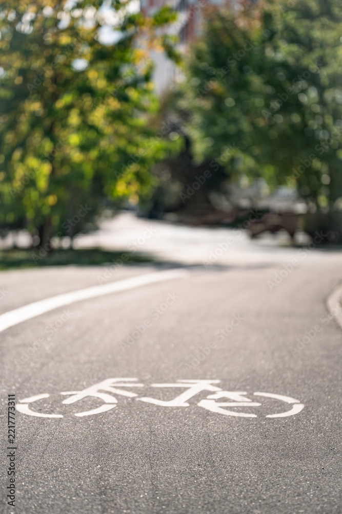 Obraz premium White bicycle road marking on an empty urban bike lane surrounded by green trees, sustainable mobility and cycling infrastructure with copy space. Vertical format.