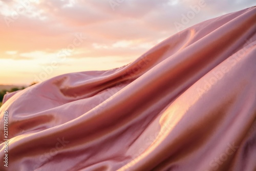 Tela de satén rosa rubor ondeando contra un suave cielo atardecer color melocotón; movimiento grácil similar a las olas y pliegues lisos y brillantes, bajo una suave luz dorada