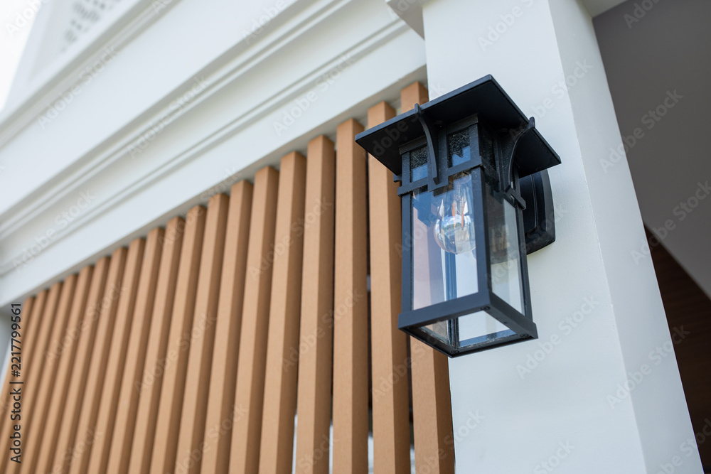 Fototapeta premium Black vintage style wall lantern with glass panels and filament bulb mounted on white pillar of modern house, wooden slats behind, exterior lighting, outdoor decor, low angle view