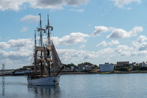 majestic vintage three masted wooden tall ship navigating through calm water near the coast on a sunny day with fluffy white clouds in the sky.