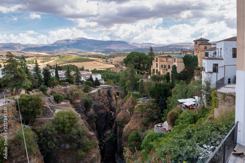 wide high angle view of el tajo canyon with historic white houses on steep cliffs surrounded by lush green foliage and distant mountains under a cloudy sky in ronda, andalusia, spain.