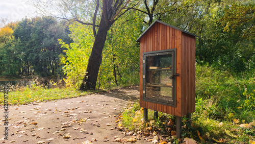 A small wooden library stands beside a walking path in a park. The library has a glass front and is surrounded by trees and fallen leaves. It is a sunny day in autumn.