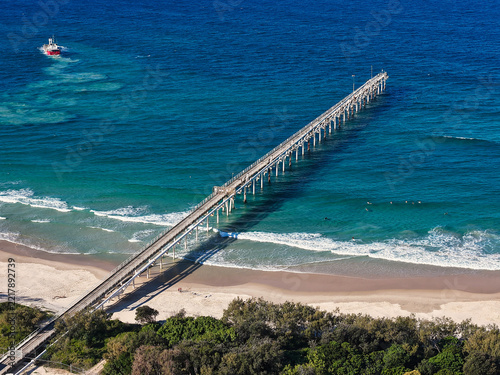 Aerial view of Cook Island pier extending into the turquoise ocean with a boat nearby and sandy beach in New South Wales, Australia.