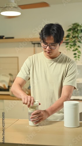 Asian man in t shirt shakes a protein shaker at home kitchen counter.
