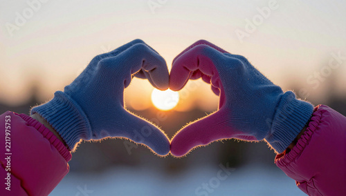 Hands in mittens form a heart around the sunset.