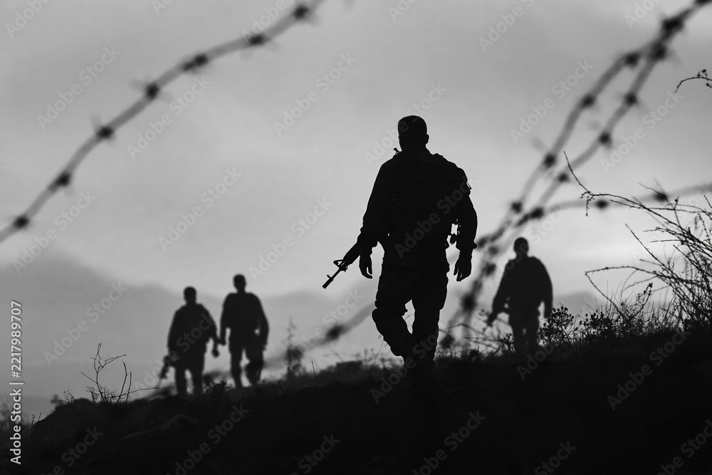 Fototapeta premium Soldiers walk along a rough landscape with silhouettes visible against a cloudy sky and barbed wire in the foreground