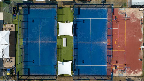 Top view of two padel courts with blue artificial turf, glass walls and white shade sails, next to a red sports court with painted lines. Zenithal shot.
