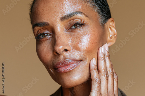 Portrait of woman gently touching cheek in warm golden light, showing calm expression and natural glowing skin against neutral background