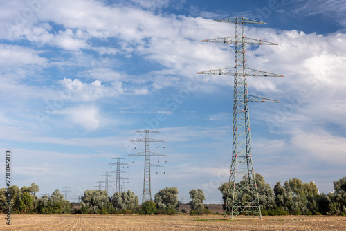 High voltage transmission tower rises at power line construction site with suspended conductors and fencing. Steel lattice structure represents grid expansion and energy engineering under cloudy sky