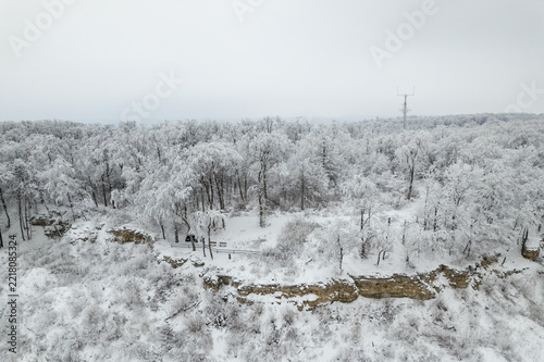 Aerial view of snow-covered forest and rocky cliff with footpath, wooden railing, and winter trees under overcast sky