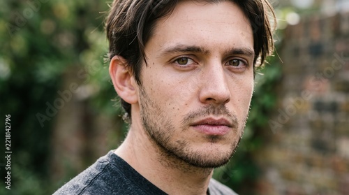 Young man gazes forward with a neutral expression outdoors against an aged brick wall.
