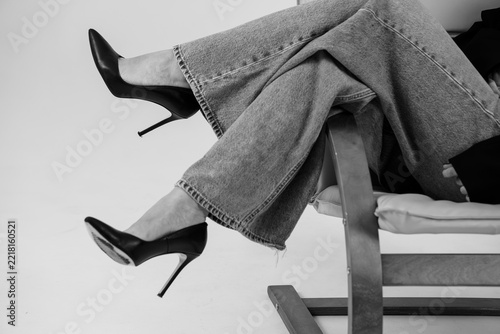 Woman's crossed legs in black heels and wide denim pants seated on a chair in black and white fashion portrait