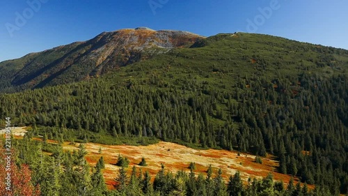 Scenic wide shot of the Babia Gora mountain range, showcasing colorful slopes with autumn vegetation, dense green coniferous woods, and a clear blue sky. 
