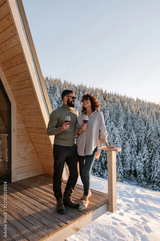 Fototapeta premium Couple enjoying wine on a snowy cabin balcony with pine forest view for winter vacation lifestyle banner and cozy holiday background