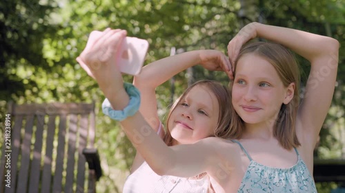 Two smiling sisters take selfie outdoors using smartphone while forming heart shape with their hands. Happy girls sit in sunny garden, sharing joyful moment during summer.
