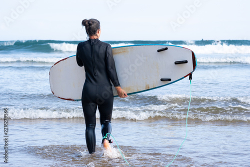Fotografie A woman in a black wetsuit walks into the sea carrying a surfboard with its fins facing the camera