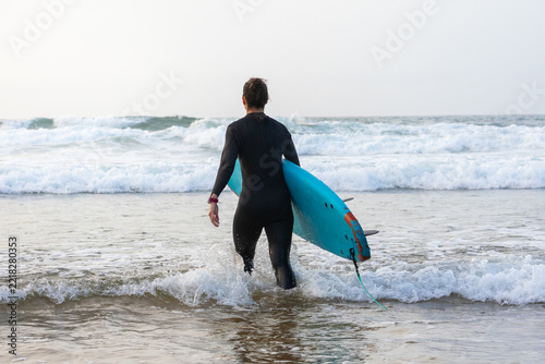 Fotografie A woman in a black wetsuit walks into breaking waves carrying a blue surfboard, viewed from behind, at the start of a surf session