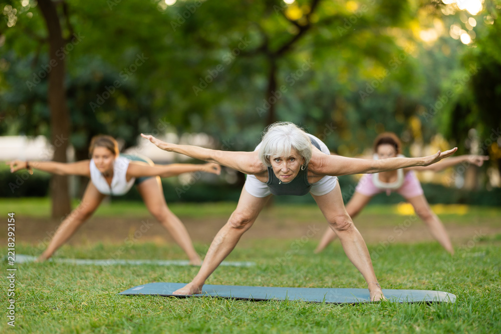 Fototapeta premium Motivated fit elderly woman participating in outdoor yoga session in green city park on summer day with group of female friends, enjoying healthy and active lifestyle