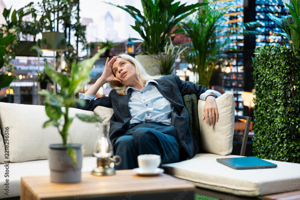 Fototapeta premium Fatigued businesswoman reclines with one hand on forehead during break, appearing overwhelmed with closed laptop beside her; relaxing on urban rooftop terrace.