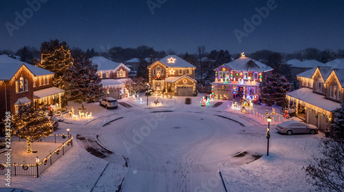 Snow-covered suburban cul-de-sac with homes decorated in holiday lights and trees. Winter neighborhood at night. Christmas and New Year celebrations. Residential holiday displays.