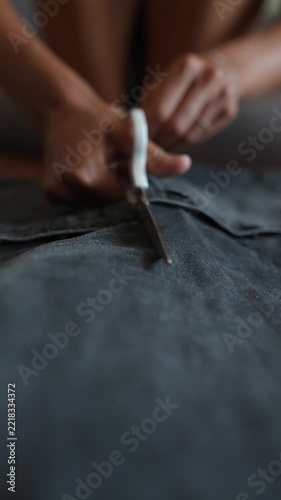 Low angle closeup of hands cutting denim seam with white handled scissors on flat work surface, showing blade separation along dark blue stitched fabric texture