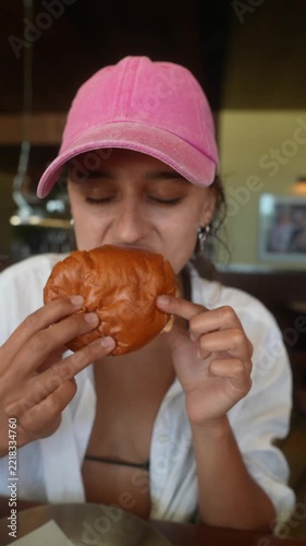 Young woman in pink baseball cap tasting burger with thoughtful side glance, indoor dining close-up, warm soft light, shallow depth of field