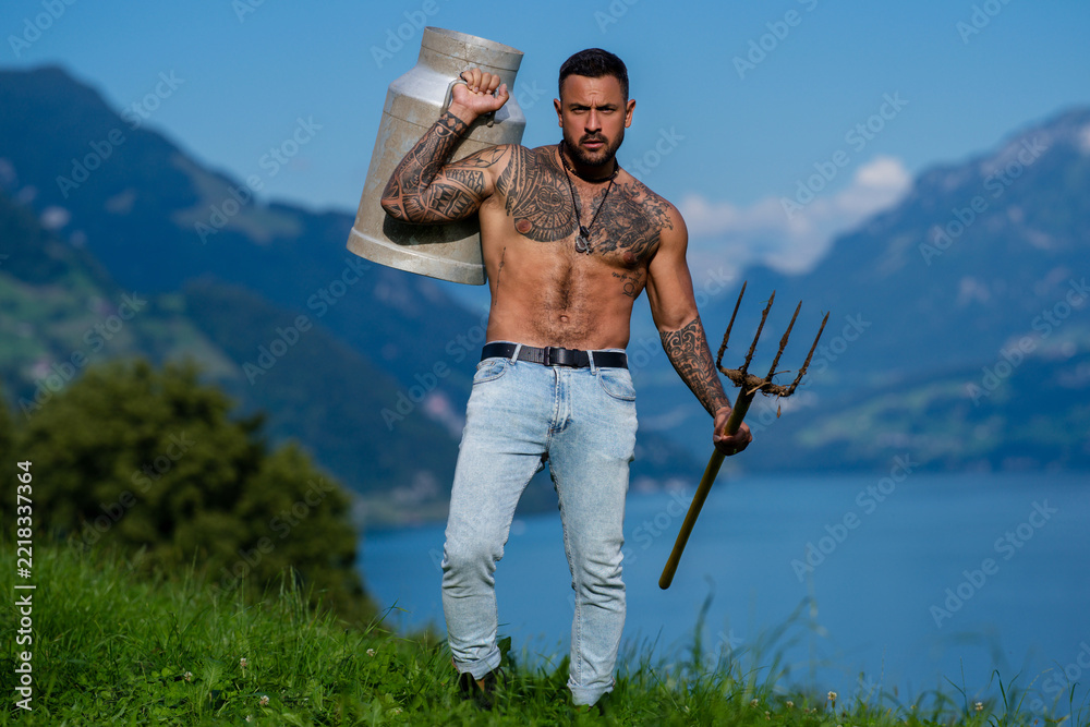 Fototapeta premium Muscular sexy farmer carrying a weathered milk can. Shirtless muscular man with visible chest tattoos. Tattooed muscular torso in a rural portrait. Muscular male model with a traditional pitchfork.