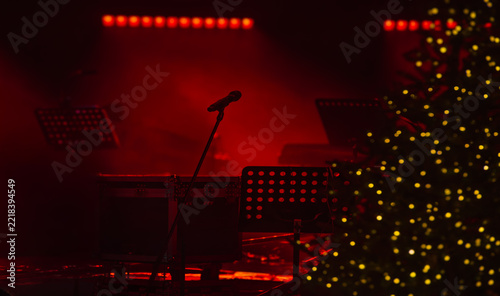 A blurred, festive concert background featuring an empty stage, red curtains, spotlight beams, and concert equipment.