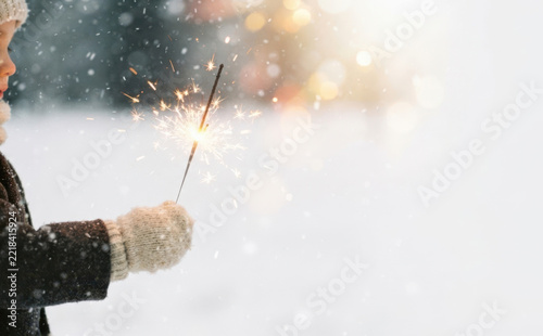 Young child holding a glowing sparkler in winter snow with festive bokeh lights for holiday celebration