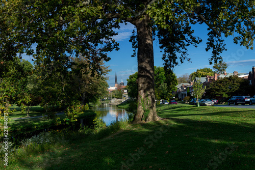 A tree-lined green park surrounds a calm waterway in Frederick, Maryland, with residential streets and church spires visible in the distance. 
