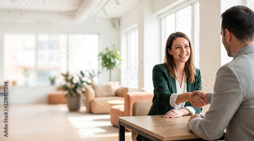 Smiling businesswoman shaking hands with a male client across a wooden desk in a bright modern office