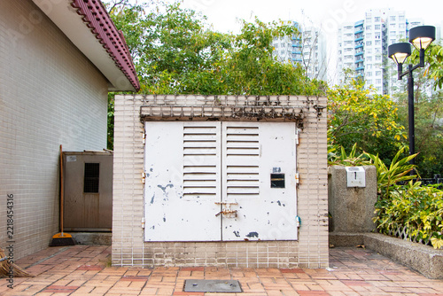 Utility substation and electrical control room located in landscaped common area of modern residential community, featuring compact utility enclosure with ventilation louvers, electrical equipment.