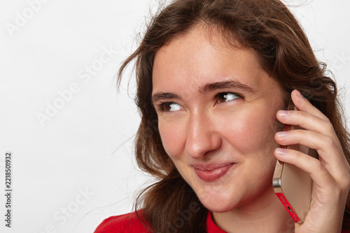 Insider promo deal, young woman with long brown hair talks on smartphone and glances slyly sideways with a cunning smile. Close-up portrait on a light background. Concept of secret deal and insider